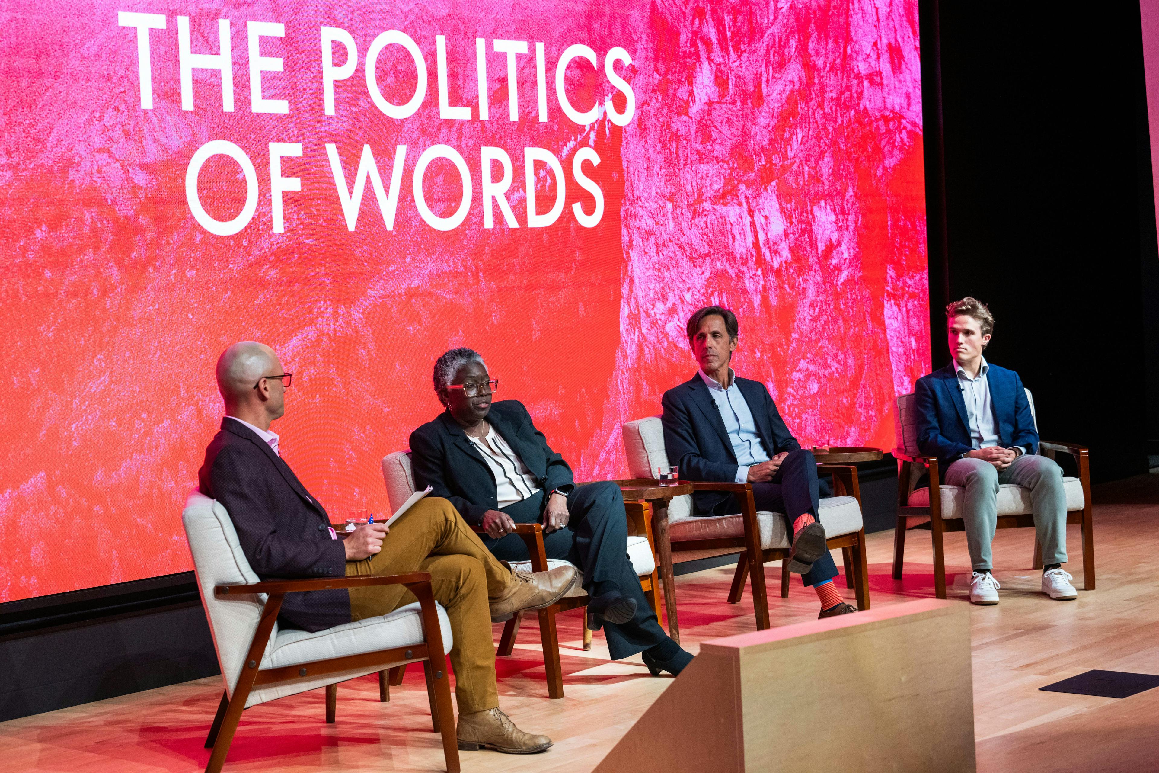 A group of 4 people sitting on chairs in front of a stage with "The Politics of Words" projected in the background