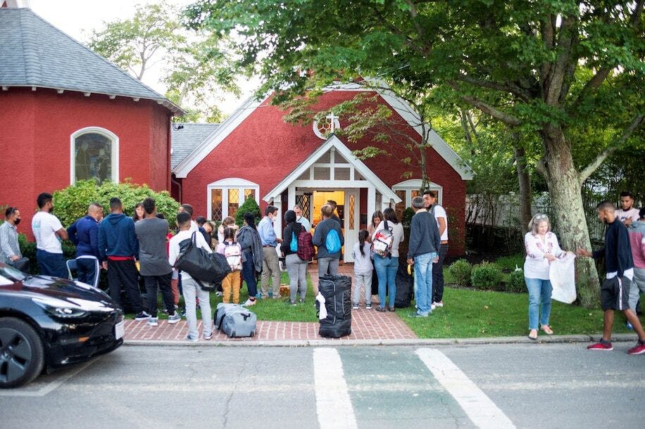 A group of a dozen people with luggages strewn about, standing in front of a red, barn-style home.